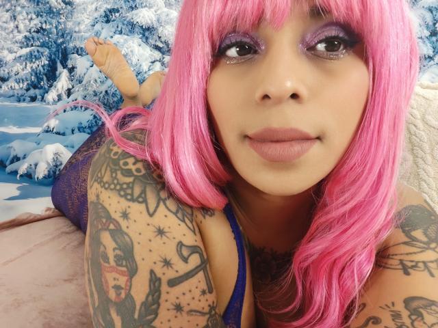 Chili with pink hair and tattoos is lying on her stomach outdoors, showing her purple eyeshadow and a slight smile.
