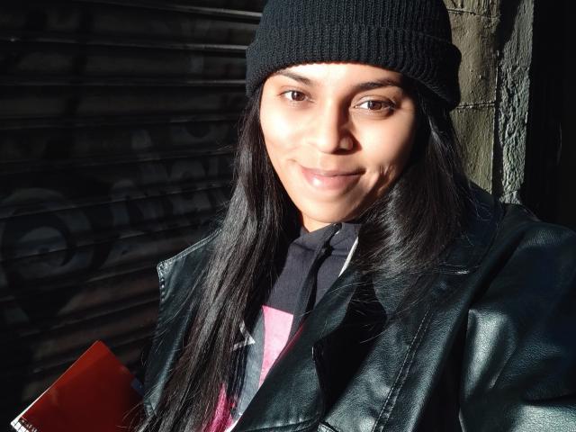 Chili, with long black hair and black beanie, smirks while wearing a black leather jacket, holding a red book.