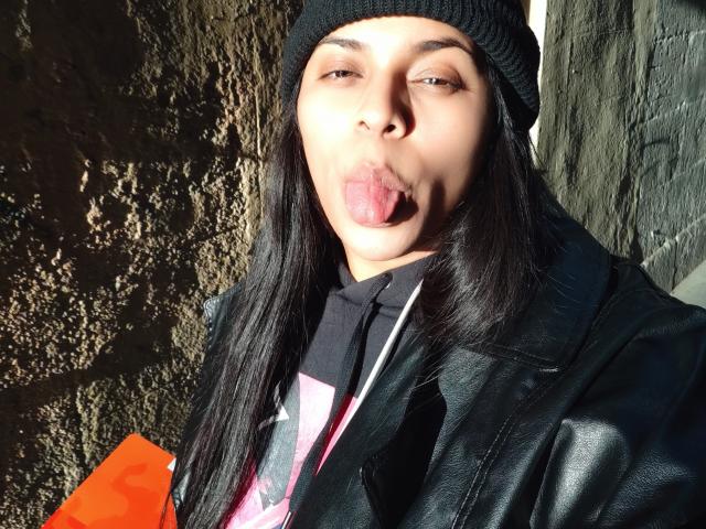 Chili is sticking out her tongue while holding an orange object against a dark, textured wall, wearing a black beanie.