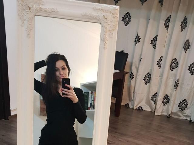 JoliDanielle with long dark hair, black long-sleeve top, small tits, holding phone in front of ornate white mirror, standing on wooden floor, smiling, arms raised.