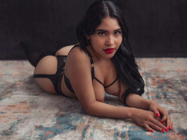 OliviGrace, avec ses cheveux noirs et son rouge à lèvres, est allongée sur un tapis en lingerie noire, avec une jarretière et des bas, montrant ses fesses et ses seins, et regardant l'appareil photo avec un sourire..