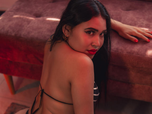 OliviGrace with long black hair, red lipstick, and earrings, wearing a black bikini, looking over her shoulder, sitting on a pink, tufted bench, red nails, slight blush on cheeks.