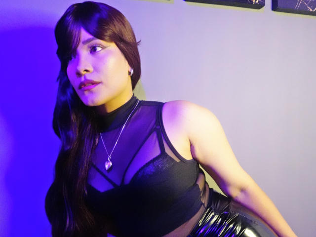 SelenaRogue, com cabelos castanhos longos, vestindo uma blusa preta transparente com amarração na frente, revelando seus seios, e calças pretas brilhantes, inclinada para frente, olhando para o lado, luz roxa..