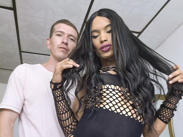 CoupleSensualHot with long black hair, purple lipstick, fishnet top, and black dress, holding hair, stands next to a white-shirted dude, both in a white ceiling room.