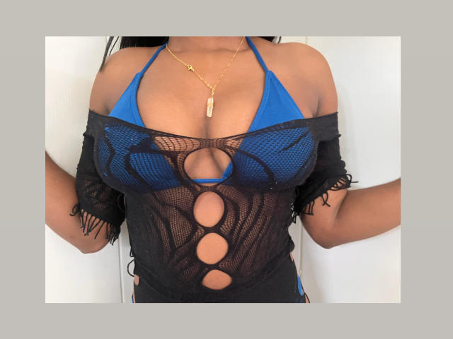 MelanyScoth con un top de bikini azul que muestra un busto grande, vistiendo una camiseta de malla negra con detalles recortados, un collar dorado y cabello largo y oscuro, de pie frente a un fondo blanco..