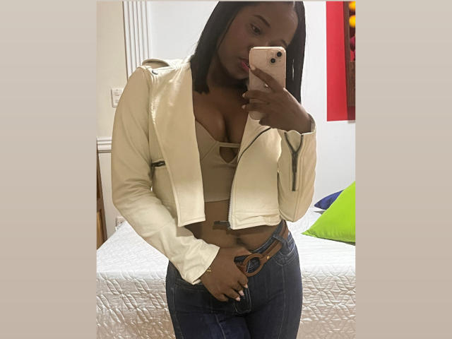 MelanyScoth with dark skin, straight black hair, wearing a white leather jacket, beige bra, and blue jeans, holding a pink phone, showing her tits and belly tattoo.