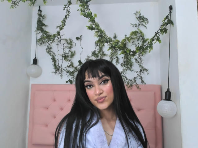 MeliMuller with long black hair, bangs, and a nose ring, wearing a white top, standing in front of a pink tufted headboard, flanked by hanging light bulbs.