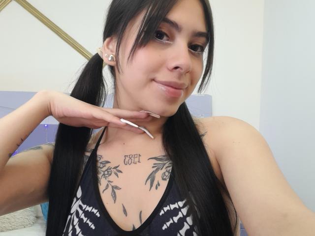 ChanelDeRosee com maria-chiquinhas, cabelo preto e tatuagens no peito, vestindo uma blusa com estampa preta e branca, sorrindo para a câmera, com a mão perto do queixo..