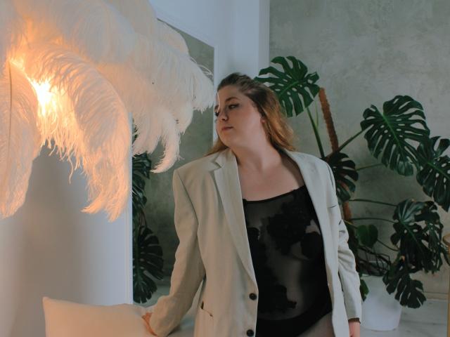 Jessika Daniels, de piel clara, cabello rubio, vestida con un blazer blanco y una blusa negra transparente, mirando una lámpara blanca con plumas, con una planta de monstera verde en el fondo..