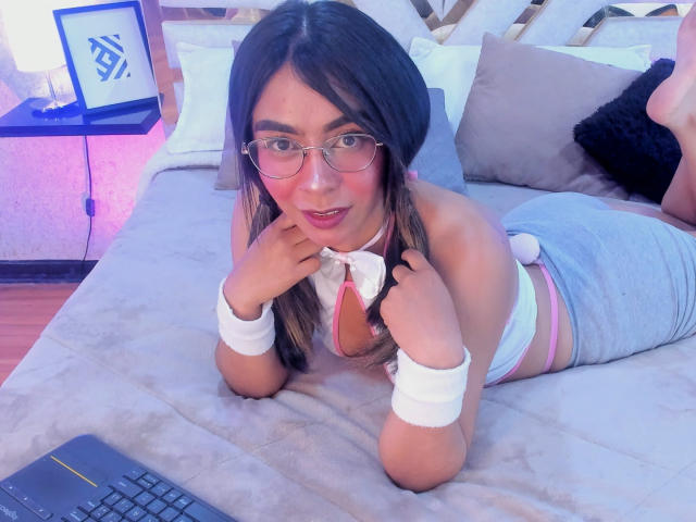 ZafiLeione avec des lunettes, un blush rose, des bracelets blancs, un débardeur gris à bordure rose, un short gris, allongée sur un lit, les mains près du visage, un ordinateur portable devant elle, dans une chambre, adorable..