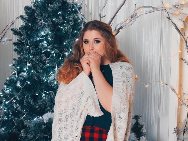 Jessika Daniels, met golvend roodbruin haar, draagt een witte gebreide trui, een zwarte top en een rode geruite rok, en staat bij een versierde kerstboom, waarbij ze aan haar vinger knabbelt..
