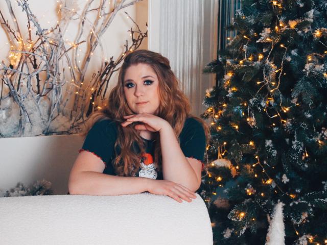 Jessika Daniels, met lang rood haar, een zwart shirt en een frons, leunt haar kin op haar hand, naast een kerstboom met witte sneeuw en gele lichtjes..