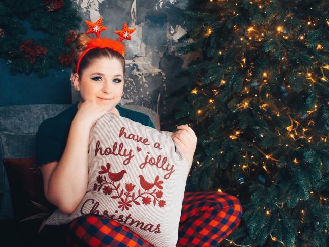 JessikaDaniels with red headband, plaid pants, and Christmas pillow, holding it against her tits, standing by a decorated Christmas tree, looking cute and slightly smug.