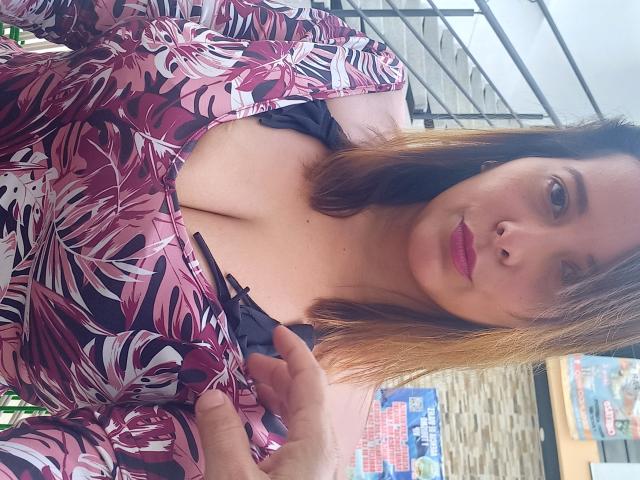 SaritaVelez with medium brown tits, wearing a pink and purple floral dress, holding black straps, pink lips, light brown hair, standing in a well-lit outdoor area.