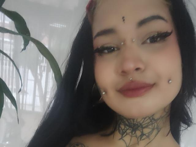 SayuriRose, con capelli neri, piercing al setto nasale e al labbro inferiore, tatuaggio a forma di ragnatela sul collo, eyeliner scuro, rossetto rosso e un leggero sorriso, in piedi accanto a una pianta verde..