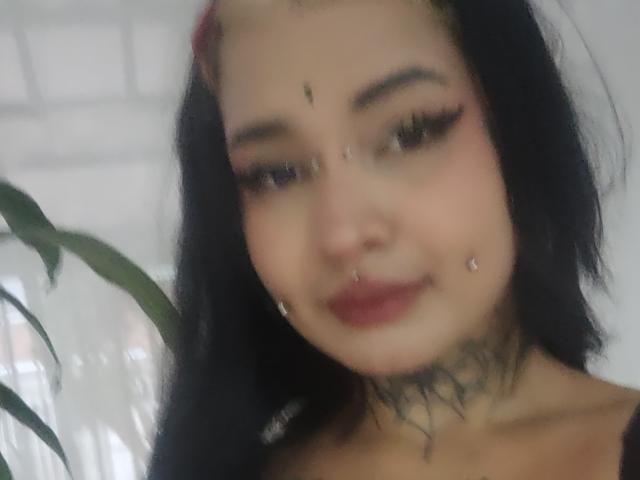 SayuriRose tem cabelo preto, sombra rosa nos olhos e batom vermelho, com piercings no nariz e no queixo, e uma tatuagem abaixo do queixo. Ela está sorrindo, com os seios quase imperceptíveis..