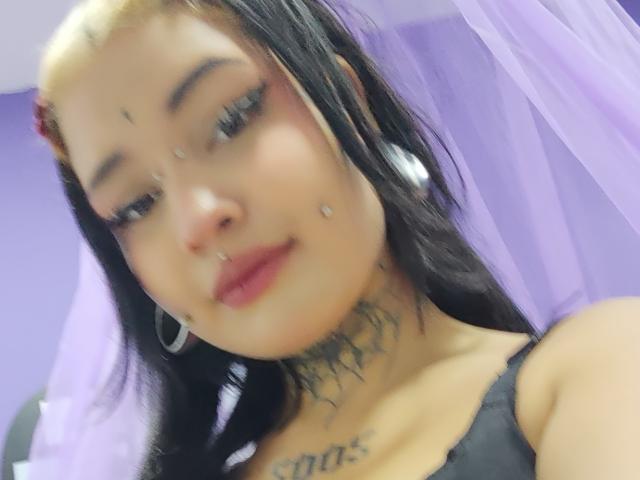 SayuriRose has black hair, a nose ring, lip ring, and multiple facial piercings. She's wearing a black top, showing tattoos on her neck and chest. Soft smile.