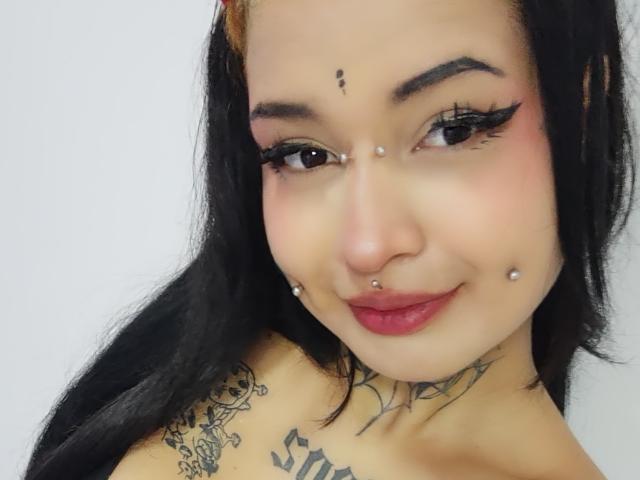 SayuriRose, avec ses cheveux noirs, ses piercings faciaux et ses tatouages, a un sourire charmant, du rouge à lèvres rouge et du crayon pour les yeux, mettant en valeur ses piercings au nez et aux lèvres, avec ses seins partiellement visibles..