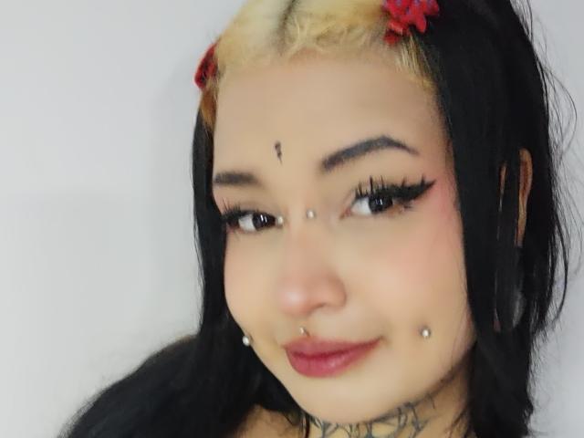 SayuriRose tiene cabello negro y rubio con flores rojas, delineador de ojos y pestañas largas. Está sonriendo y tiene un piercing en la nariz, un piercing en el labio y piercings faciales. Sus pechos son pequeños..