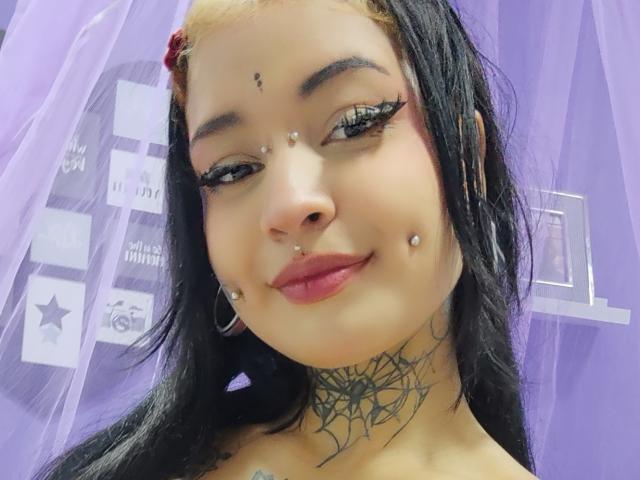 SayuriRose, con cabello negro, mechas rubias y piercings en la nariz, las mejillas y el labio superior. Tiene un tatuaje de telaraña en el cuello y lleva pintalabios rosa..