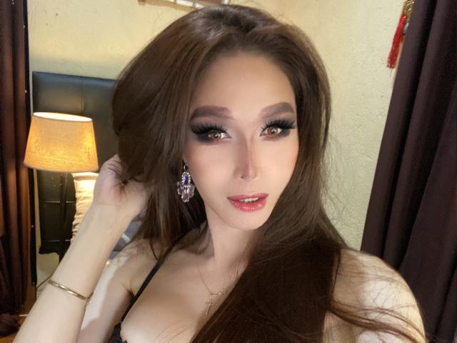 LaurentSuprema with long brown hair, dark eyeliner, pink lipstick, small earrings, black top, hand in hair, looking at camera, slight smile, soft light, warm skin, small tits.
