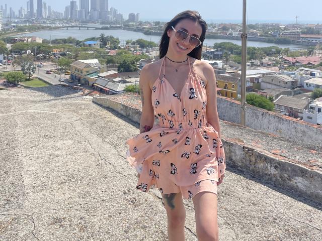 LeilaP, con un vestido rosa con un estampado de mariposas, lo sostiene extendido, mostrando un tatuaje en su muslo, usando gafas de sol y un collar negro, de pie en una azotea de grava..