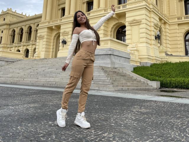MeganWisse stands with arm raised, smiling. She's wearing a white off-shoulder top, tan cargo pants, and white sneakers. Her long brown hair is down. She's outside a yellow building.