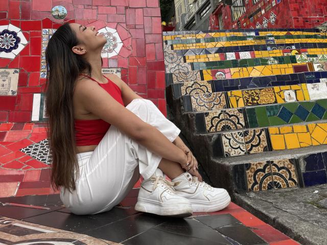 MeganWisse with long brown hair, red crop top, white pants, and white sneakers, sitting on colorful tile stairs, head tilted back, hands on knees, looking up.
