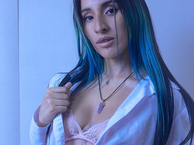 LeilaP, com cabelo azul-esverdeado, piercing no nariz e no lábio, vestindo uma camisa branca aberta para revelar seios femininos cor-de-rosa com renda, segurando a camisa, usando um colar roxo, com iluminação azul..