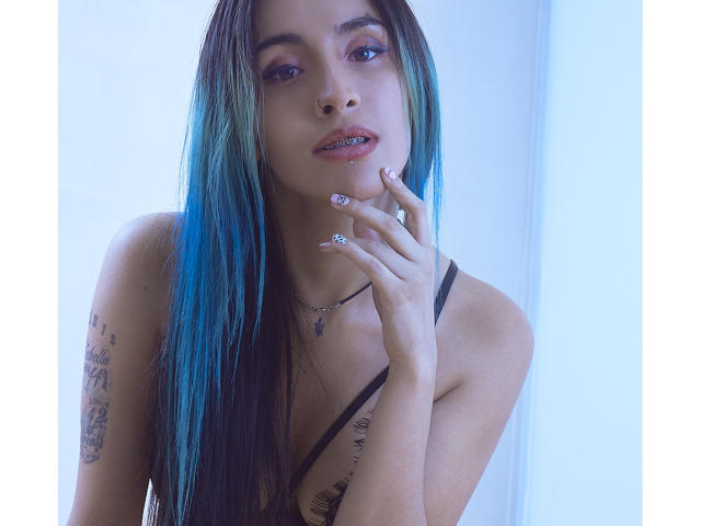 LeilaP, con cabello largo azul verdoso, un piercing en la nariz y un anillo en el dedo, se toca la barbilla, mostrando tatuajes en su brazo izquierdo, lleva una correa negra y tiene una expresión seductora..