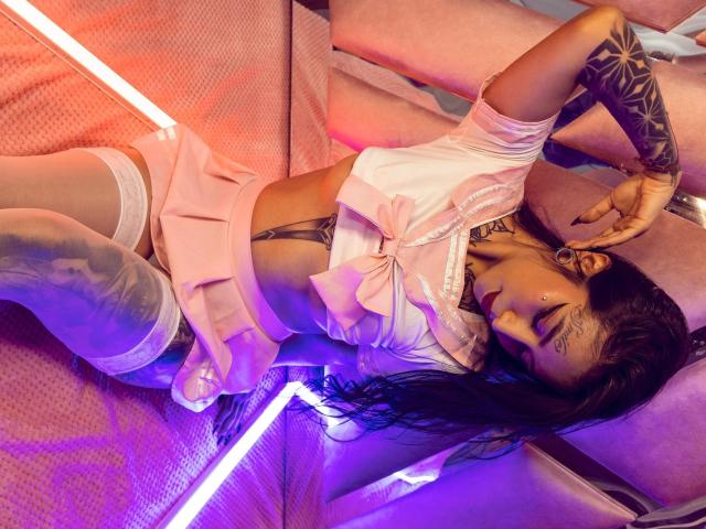 KarolyLove, lying on pink couch, wearing pink frilled top, white garter, sheer thigh-highs, tattooed arm, black hair, purple neon light, hands near face, legs spread.