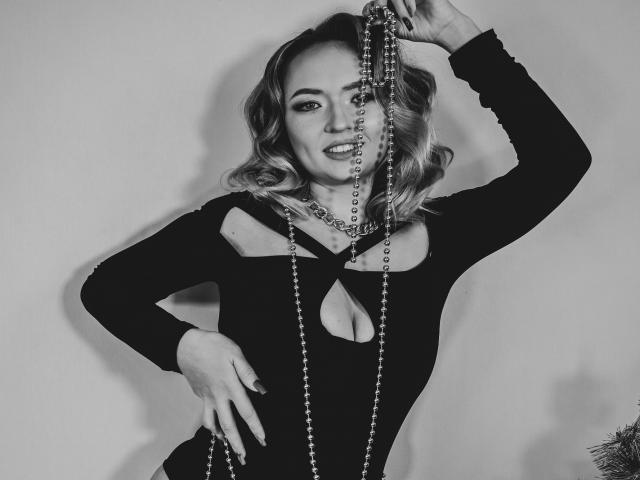 SallyVerda with wavy blonde hair, wearing a black cutout dress, holding bead necklaces, smiling, one arm raised, other hand on hip, gray-black photo, sexy, cute, confident.