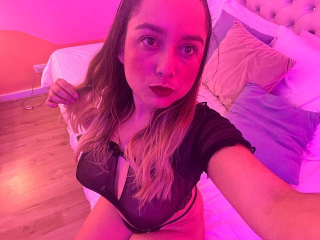 AmayaJhons dans une chambre éclairée en rose et violet, portant un haut noir transparent qui dévoile sa poitrine, avec de longs cheveux blonds, assise sur le lit, montrant ses seins et son culotte rayée..