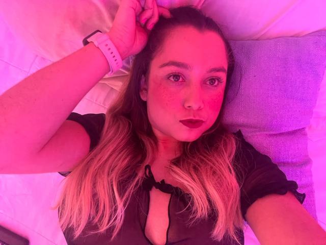 AmayaJhons with pink-tinted skin, long blonde hair, dark lipstick, black shirt with cleavage, wristwatch, lying on pillow, hand in hair, looking at camera, slightly freckled face.