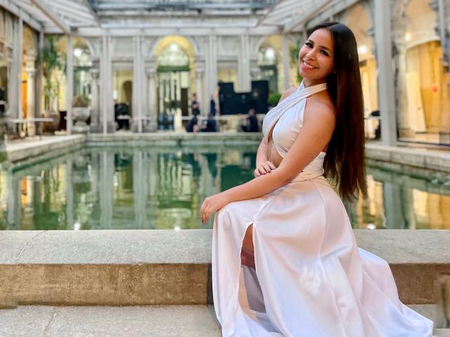 MeganWisse with long brown hair, smiling, in a white, sleeveless, high-slitted dress, sitting on a stone ledge by a reflective, indoor water pool. Boobs, ass, pretty.