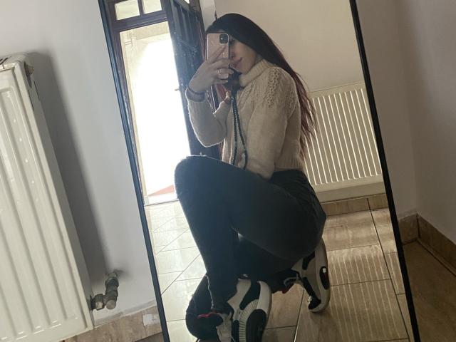 RinaQueenie squatting in front of a mirror, wearing a white knit sweater, black pants, and white Adidas sneakers, with long brown hair and holding a phone.