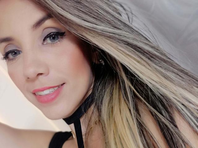 LindsaySummer heeft blond en bruin haar, roze lippen, zwarte eyeliner en een zwarte choker. Ze glimlacht met witte tanden en kijkt naar de camera. Haar borsten zijn gedeeltelijk zichtbaar..