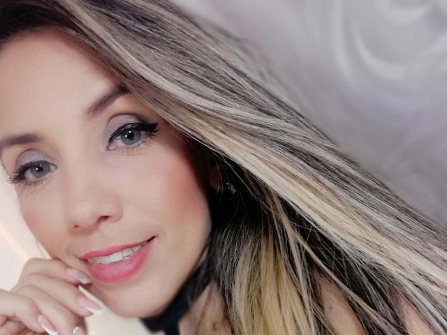 Lindsay Summer tiene el pelo largo, rubio y castaño, ojos azules con delineador negro, labios rosados y una sonrisa encantadora. Está tocándose la mejilla con la mano derecha..