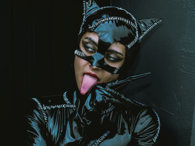 SerenaDeStark trägt glänzendes schwarzes Latex mit Katzenohren, einer Maske und hat die Zunge herausgestreckt. Ihre Augen sind geschlossen, und sie hat einen sexy, verspielten Ausdruck. Ihre Brüste sind teilweise sichtbar..