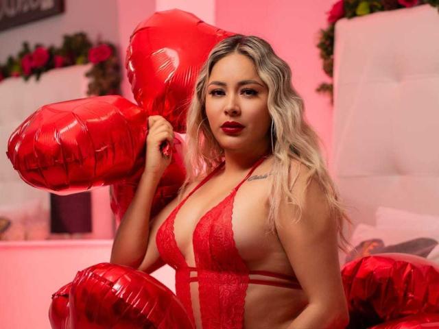 CamilaBardot with blonde hair, red lace lingerie, holding shiny red heart balloons, showing her tits, red lipstick, and a tattoo on her right shoulder, in a pink-lit room.