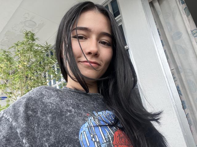 DeisyWest has long black hair, a cute face with a slight smile, and is wearing a gray graphic tee. She's standing near a window with a plant.