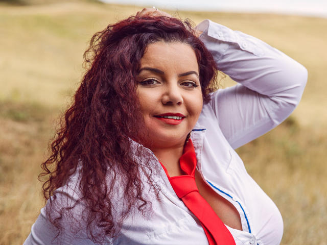 MiryamInes, con cabello rizado y pelirrojo, vestida con una camisa blanca y una corbata roja, con la mano en el cabello, sonriendo a la cámara, mostrando ligeramente sus senos a través de la camisa, al aire libre..
