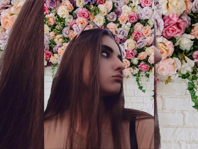 KariMillie tiene el pelo largo y castaño, una expresión pensativa, y lleva una blusa transparente. Está frente a una pared cubierta de rosas rosas, blancas y moradas..