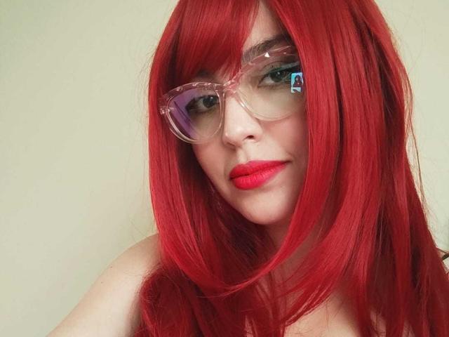 JackyEmma with bright red hair, clear glasses, and red lipstick, showing a slight smile, has pale skin and is wearing nothing visible above the chest.