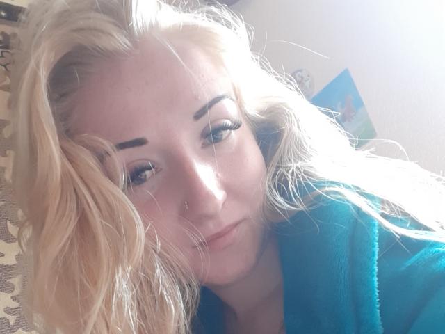 BonnyMyriam, con cabello rubio ondulado, ojos azules y un piercing en la nariz, vestida con una bata color turquesa, se ve cansada y con el ceño fruncido. Sus senos no son visibles. Está en una habitación iluminada por el sol..