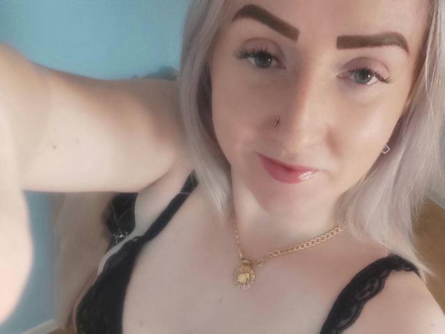 BonnyMyriam, con piel pálida, cabello rubio platino, cejas oscuras, piercing en la nariz, sostén de encaje negro, labios rosados y brillantes, collar dorado, sonriendo, selfie, fondo azul, maquillaje sutil..