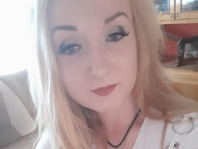BonnyMyriam has blonde hair, dark eyebrows, glittery green eyeshadow, thick black eyeliner, red lipstick, nose piercing, white shirt, black necklace, and a slight smile.