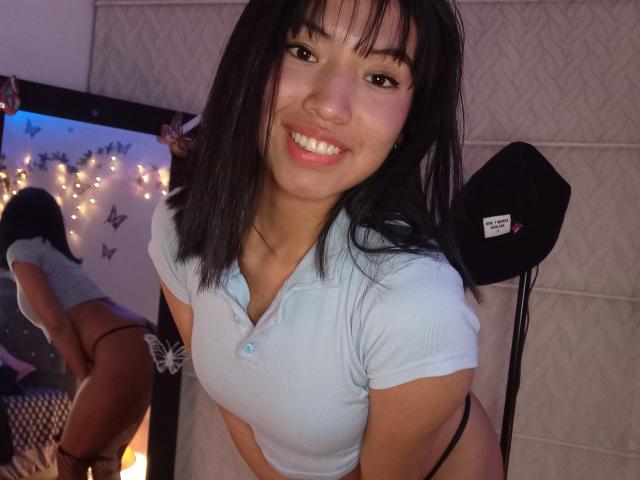 Lucia Garcia, con i capelli neri, sorridente, indossa una camicia azzurra, mostrando il sedere in un riflesso allo specchio, con uno slip nero e un cappello nero su una sedia..