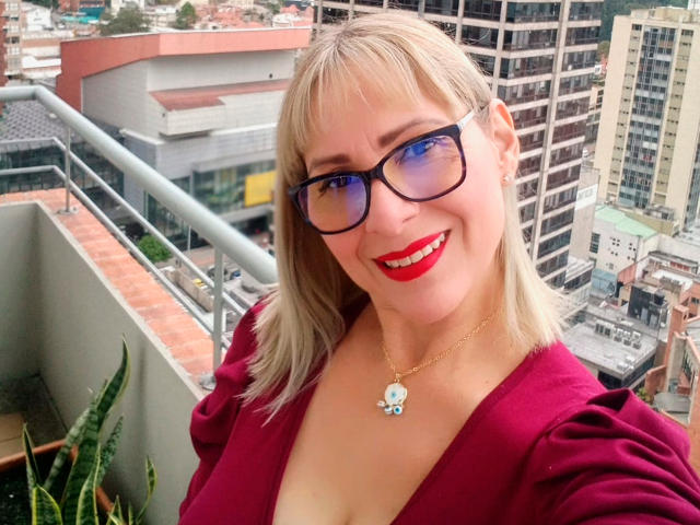 Jane Turner, met blond haar, een blauwe bril, rode lippenstift en een rode jurk, laat haar borsten zien, glimlacht en draagt een ketting met een blauwe steen. Op de achtergrond een stedelijk balkon..