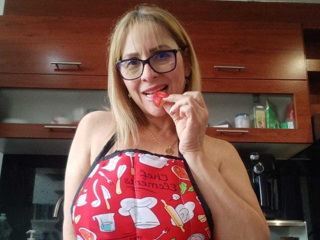 JaneTurner with blonde hair, glasses, and a red apron with kitchen patterns, biting a red strawberry, showing her tits, smiling in a kitchen.